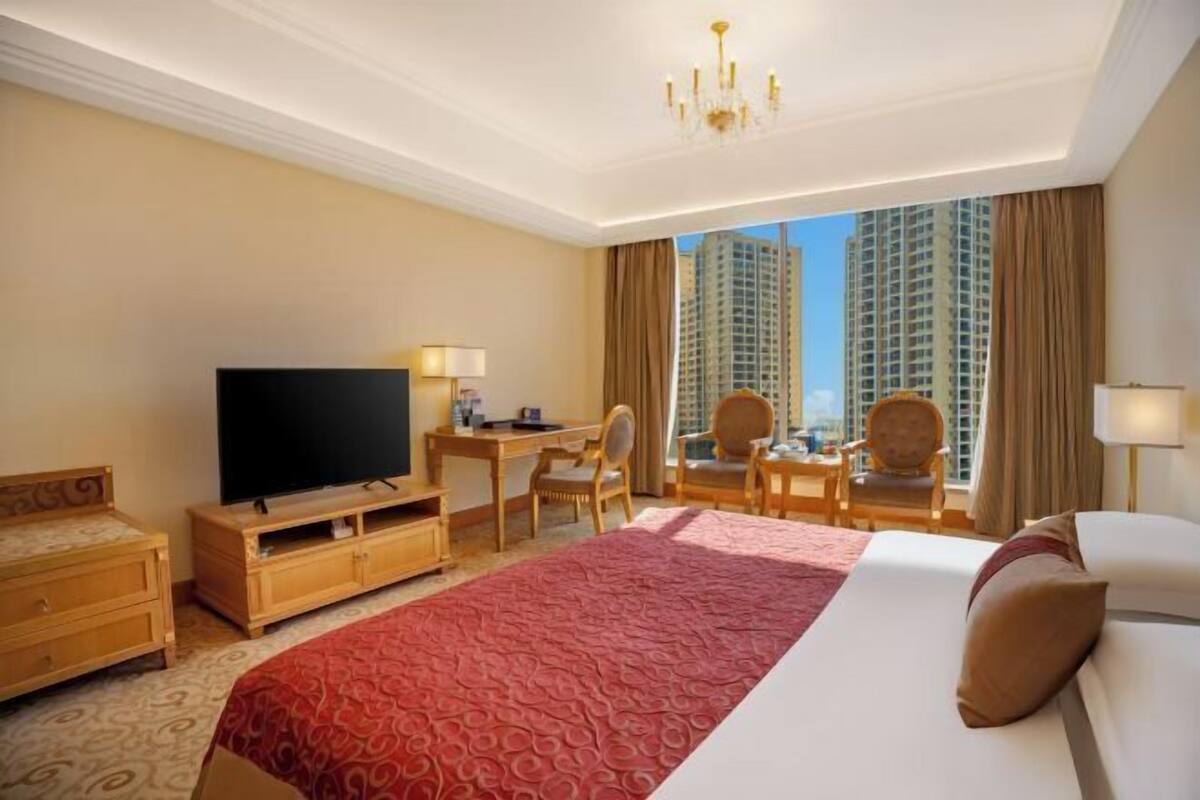 A hotel room with a large bed, a TV, a desk with chairs, and a view of tall buildings.