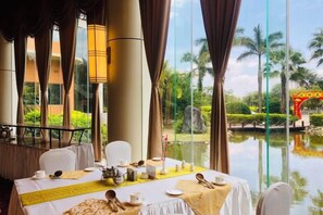 A dining area with a table set for four, large windows, and a view of a water body and greenery. - Honghe Guanfang Hotel (Mengzi)