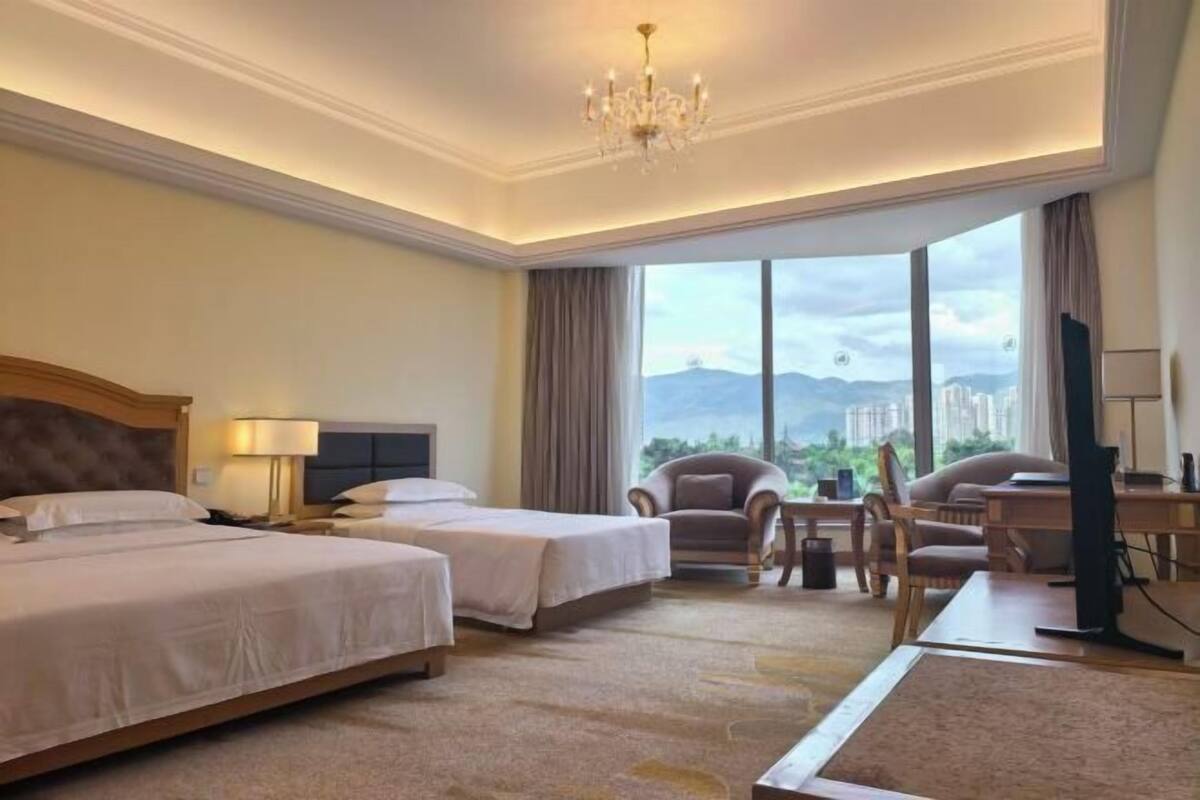 A hotel room with two beds, a desk, a chair, a TV, and a large window with a city view.