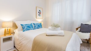 Een keurig opgemaakt slaapkamer met een bed, een nachtkastje met een lamp, een lijst met een foto aan de muur, en een raam met dunne gordijnen.