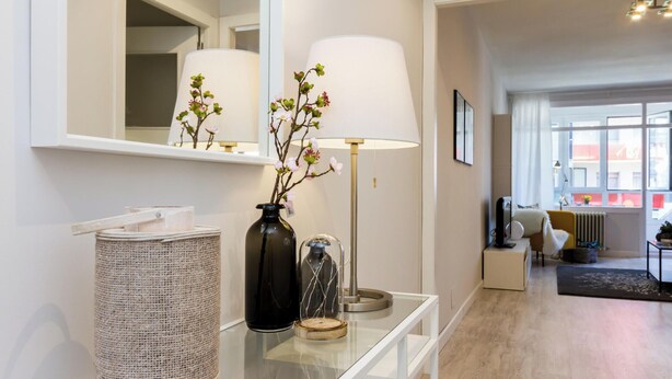 Een moderne hotelkamer met een glazen console, een lamp, een vaas met bloemen, een rieten mand en een grote spiegel die de kamer weerspiegelt.