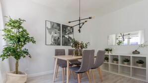 Een eetruimte met een houten tafel en grijze stoelen, een grote plant en abstracte kunst aan de muur.