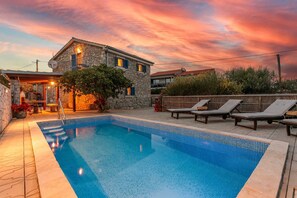 Una piscina con sillones de descanso, una casa de piedra y un cielo al atardecer.