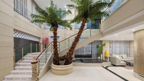 Un moderno vestíbulo de hotel con una escalera, palmeras, un sofá y una lámpara.