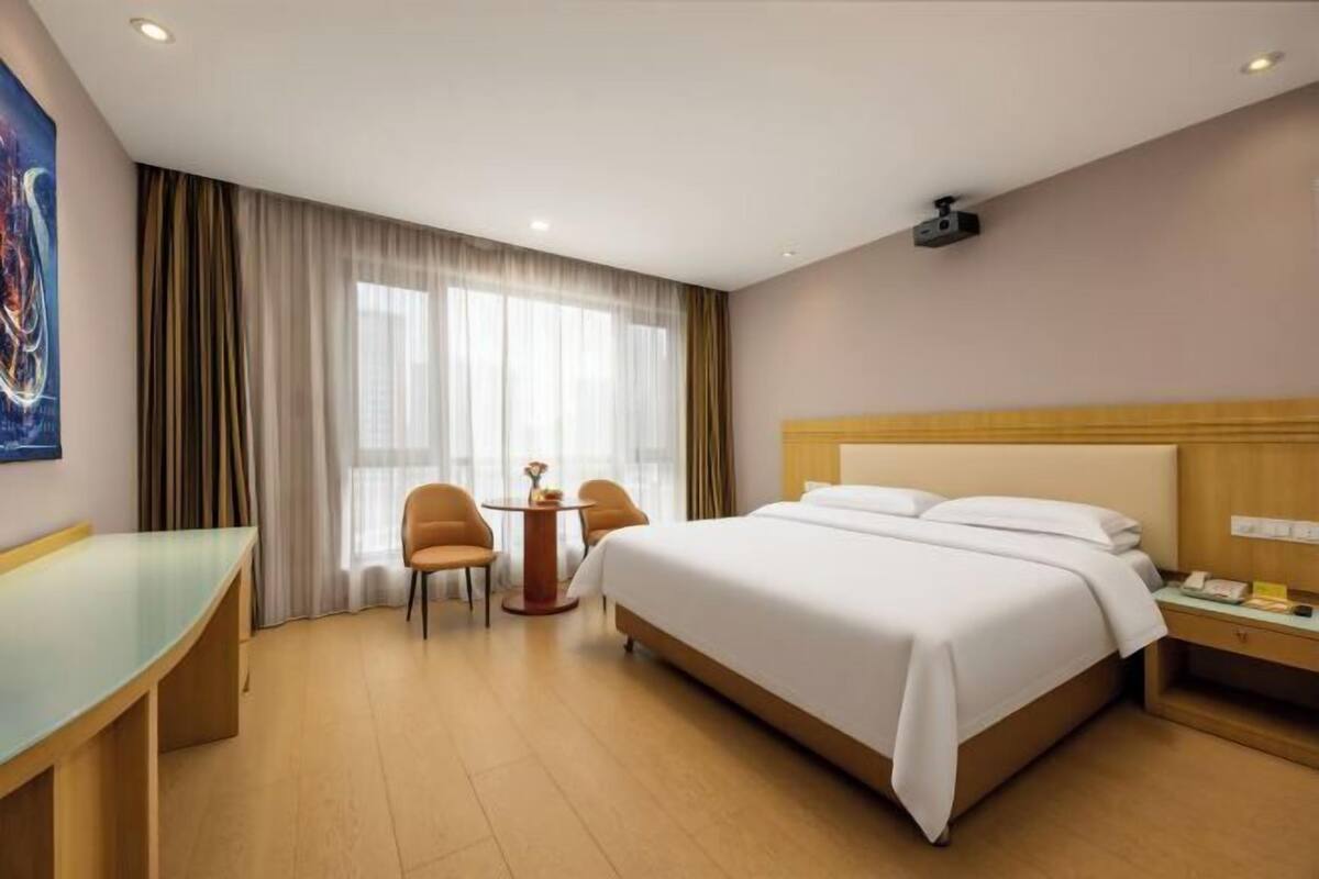 A hotel room with a bed, a desk, two chairs, a small table, and a window with curtains.