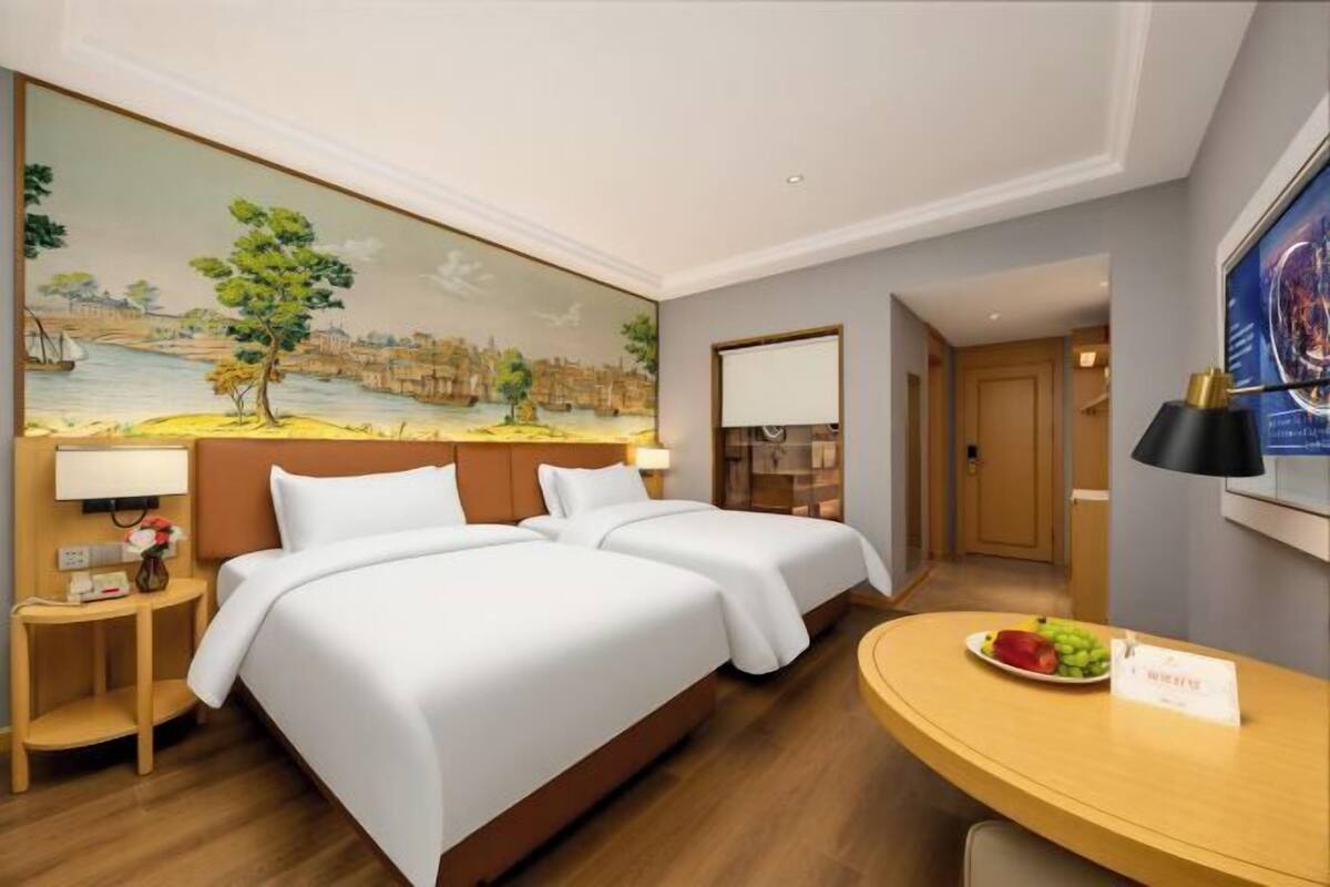 A hotel room with two beds, a painting on the wall, a desk with a fruit plate, and a view of the hallway.