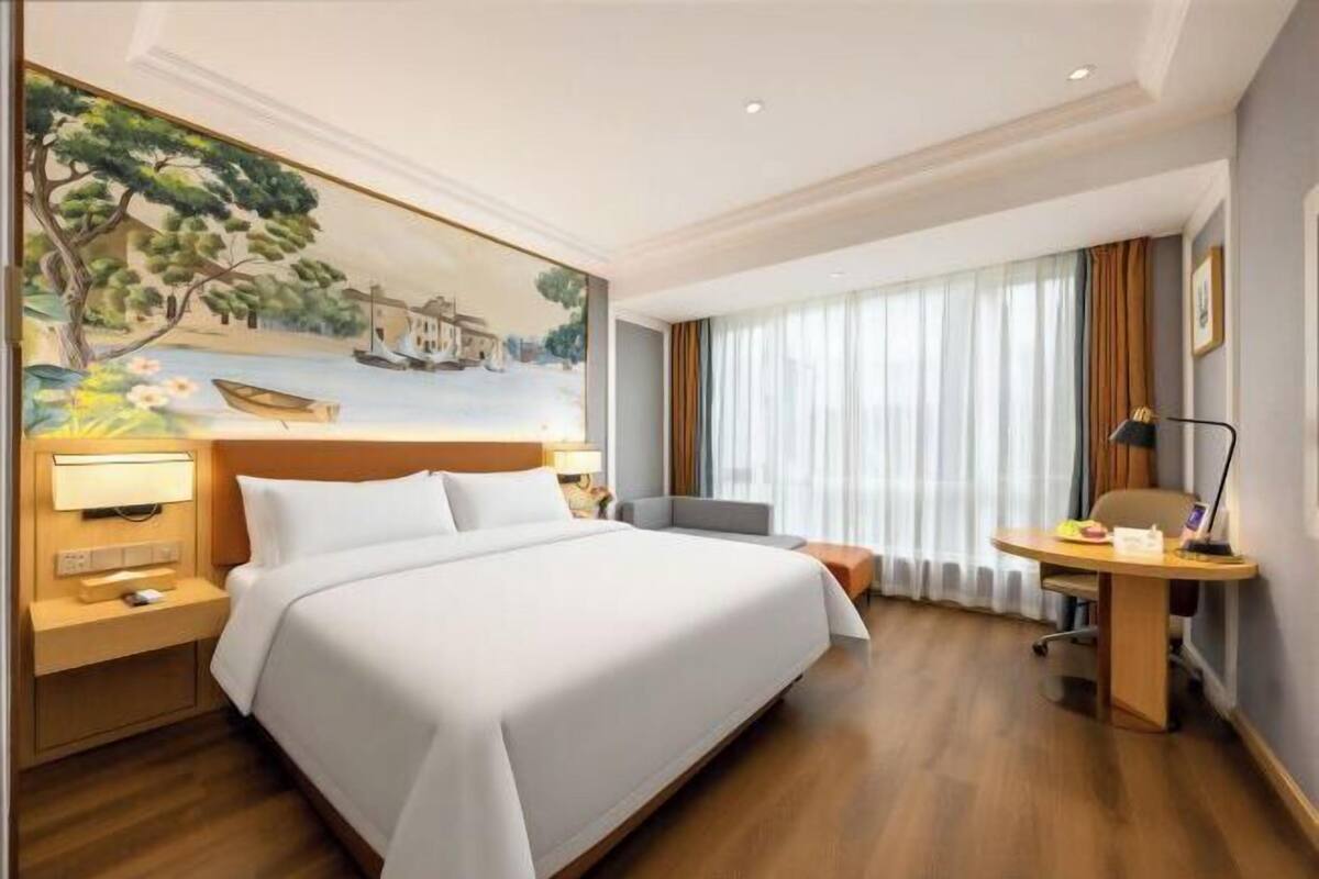 A hotel room with a bed, a desk, a chair, a nightstand, and a painting on the wall.