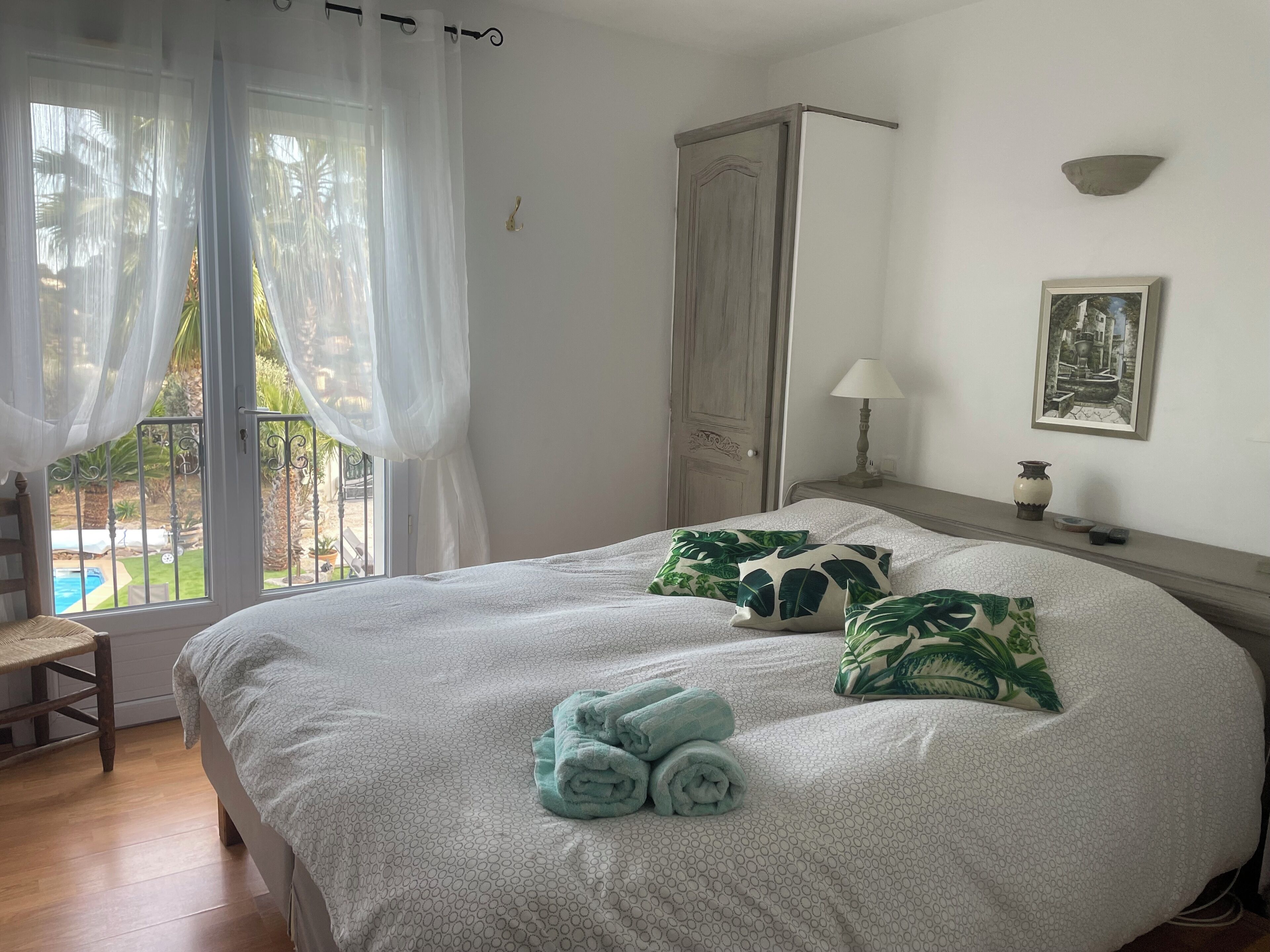 A bedroom with a bed, a nightstand, a lamp, a framed picture, and a view of a pool through sheer curtains.