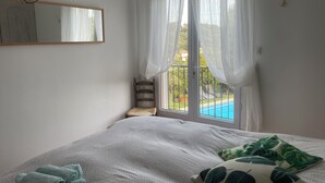 A bedroom with a bed, a TV mounted on the wall, a window with sheer curtains, and a view of a pool outside.