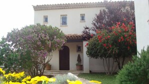 A building with a tiled roof, a central entrance, and a garden with yellow flowers and various shrubs.