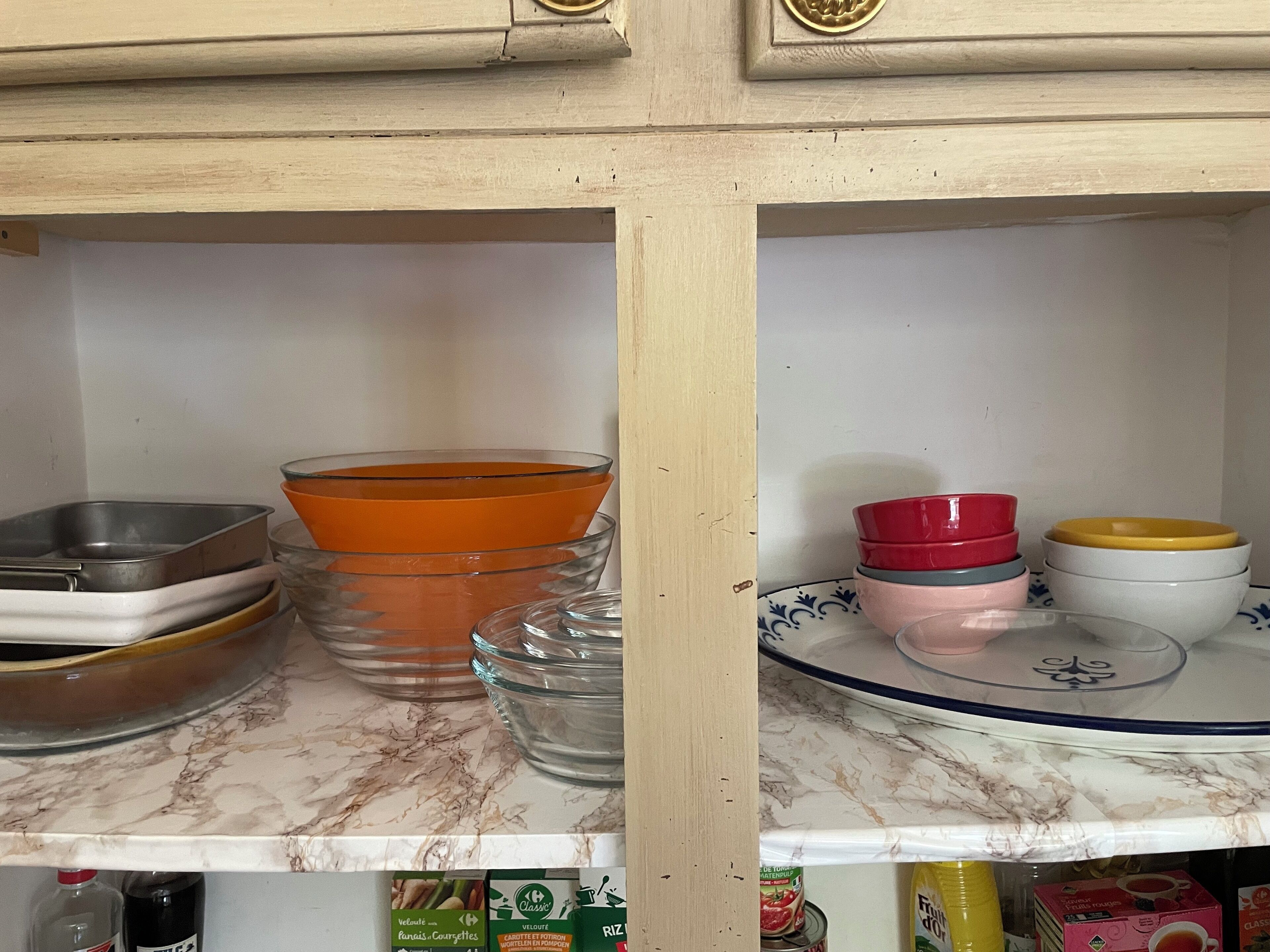 A kitchen shelf with various dishes and containers, including a metal tray, orange bowls, and a glass jar.
