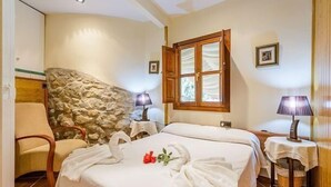Hotelkamer met een stenen muur, een bed met witte lakens, een stoel, twee nachtkastjes met lampen en een raam met houten luiken.