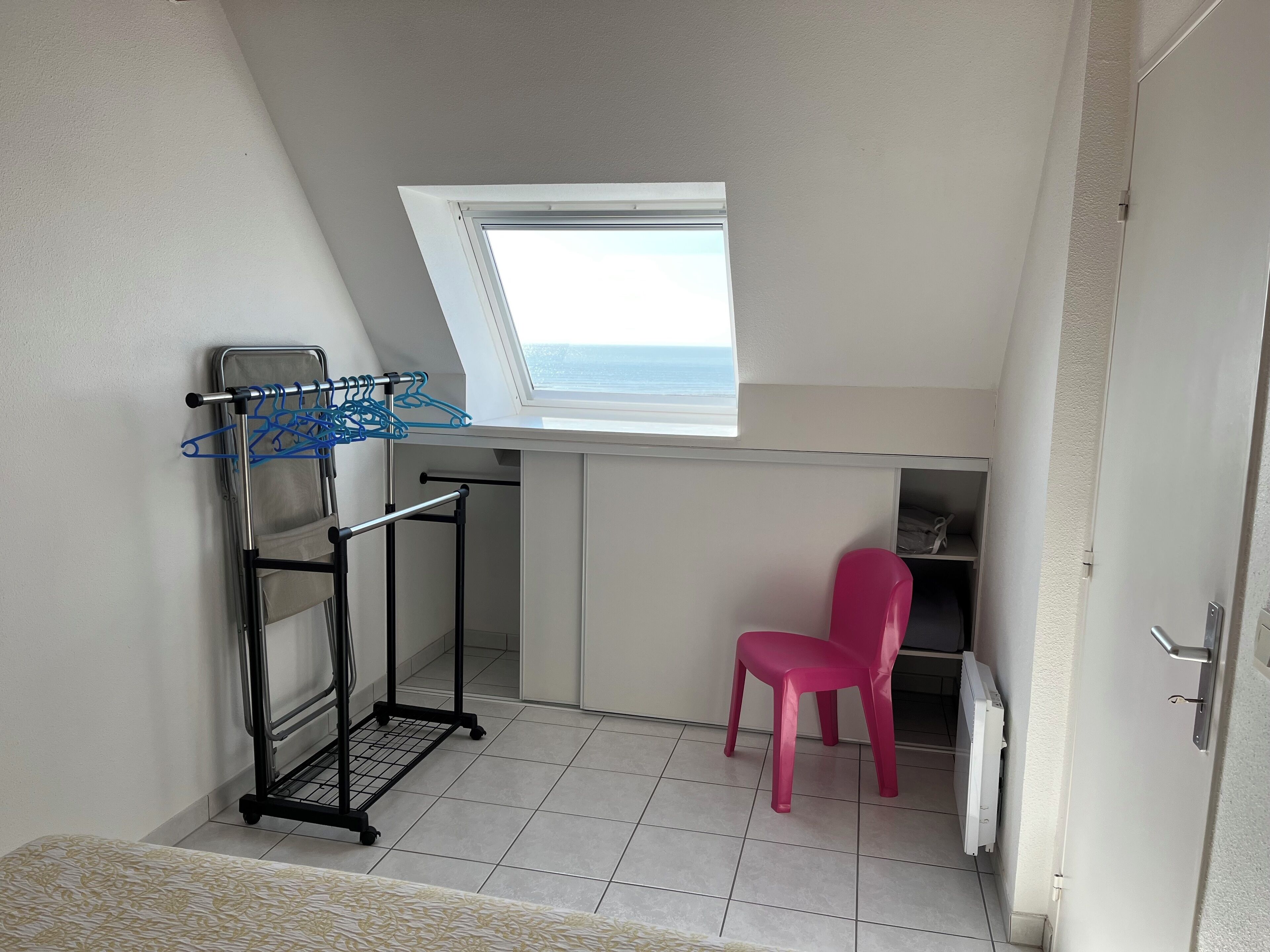 A small room with a skylight, a pink chair, a metal drying rack, and a white door.
