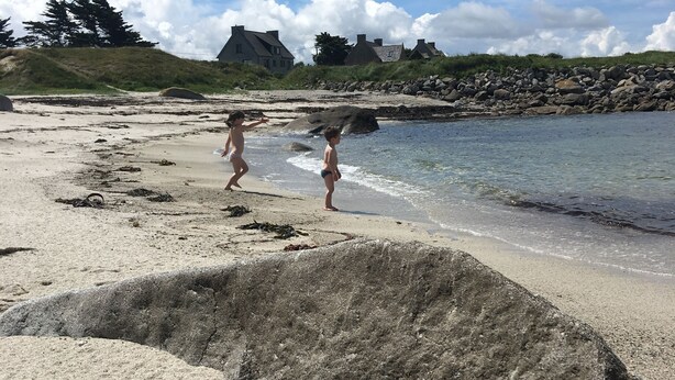Ein Sandstrand mit Fußspuren, eine felsige Küstenlinie und Häuser im Hintergrund.