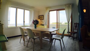 Ein Essbereich mit Holztisch und Stühlen, einem Blumenstrauß und Blick auf das Meer durch ein großes Fenster.