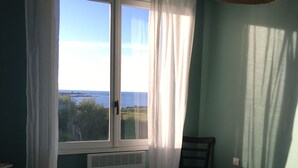Ein Schlafzimmer mit Bett, Stuhl, einem Fenster mit Vorhängen und Meerblick.