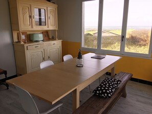 A dining area with a wooden table, white chairs, a bench with a black and white pillow, a vintage-style cabinet, and a view of the countryside through a large window. - The sea at your feet in Brittany! (Plouescat)