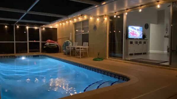 An indoor swimming pool with a tiled edge, a lounge area with chairs and a table, and a television screen in the background.