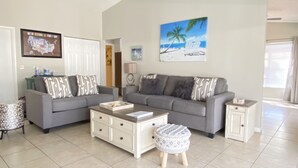 Ein Wohnzimmer mit zwei grauen Sofas, einem Couchtisch, einem gemusterten Ottomanen, einer weißen Kommode und einem Gemälde mit einer Strandansicht.
