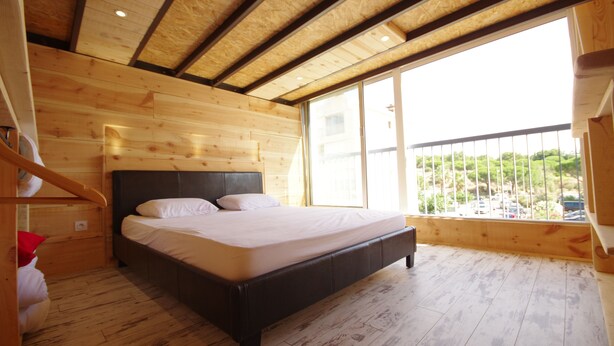 A wooden-paneled bedroom with a large bed, a balcony with a railing, and a view of greenery outside.