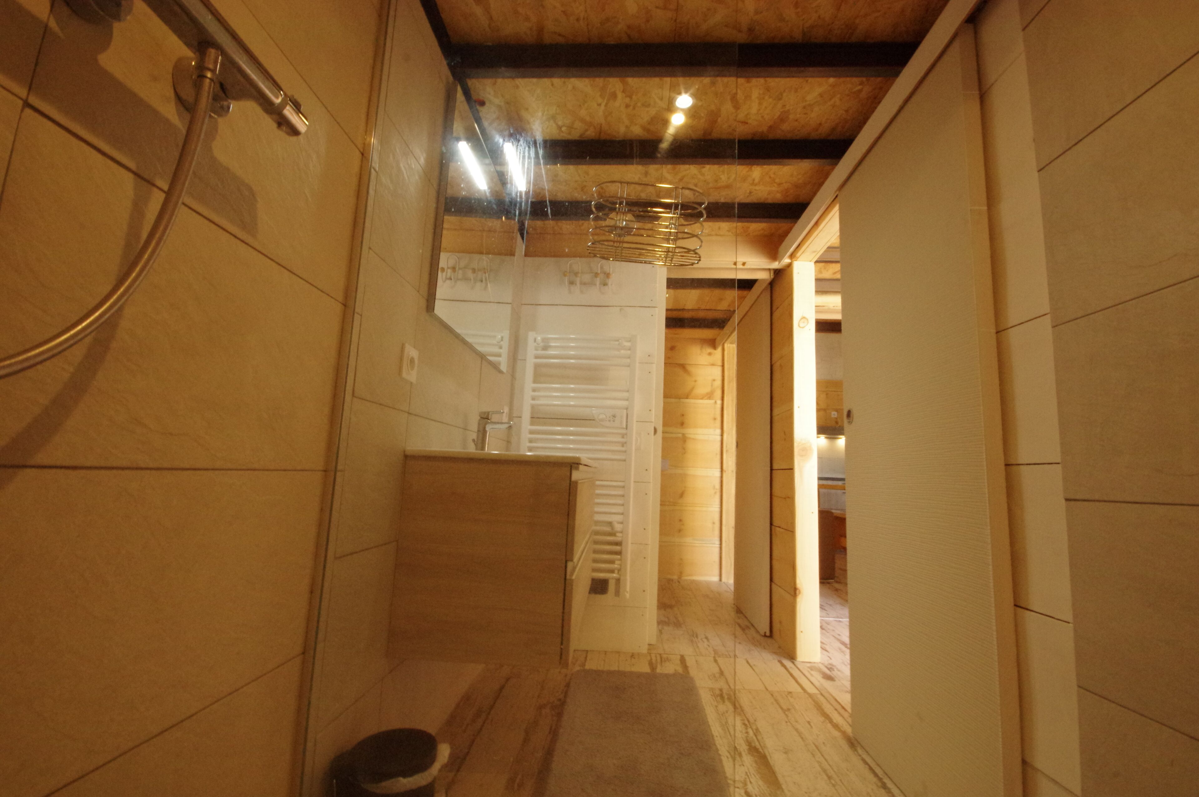 Ein schmales Badezimmer mit Fliesen, einer Dusche, Holzbalken an der Decke und einer sichtbaren Tür zu einem anderen Raum.