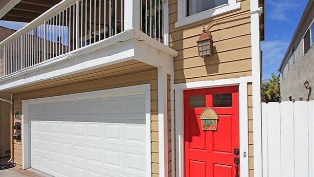 A two-story house with a white garage door, a red front door with a mailbox, and a white picket fence.