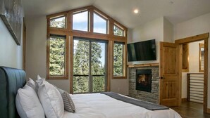 A bedroom with a large bed, a stone fireplace, a TV mounted above it, and a view of trees through large windows. - ⭐️Luxury newly built with Sauna, Steam and Hot Tub  (Truckee)