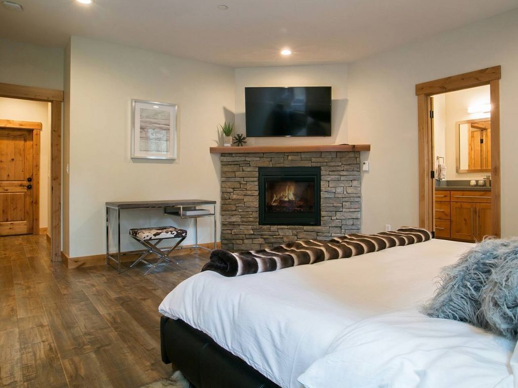 A bedroom with a stone fireplace, a bed with white bedding, a television above the fireplace, and a wooden door.