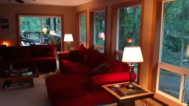 A cozy living room with a red sofa, a coffee table, and a lamp. Large windows provide a view of the outdoors. - Relax and Rejuvenate in this Peaceful, Wooded Oasis. (Eureka Springs)