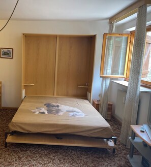 Ein Zimmer mit einem Bett, einem Holzschrank, einem kleinen Tisch, einem Spiegel und einem Fenster mit Vorhängen.
