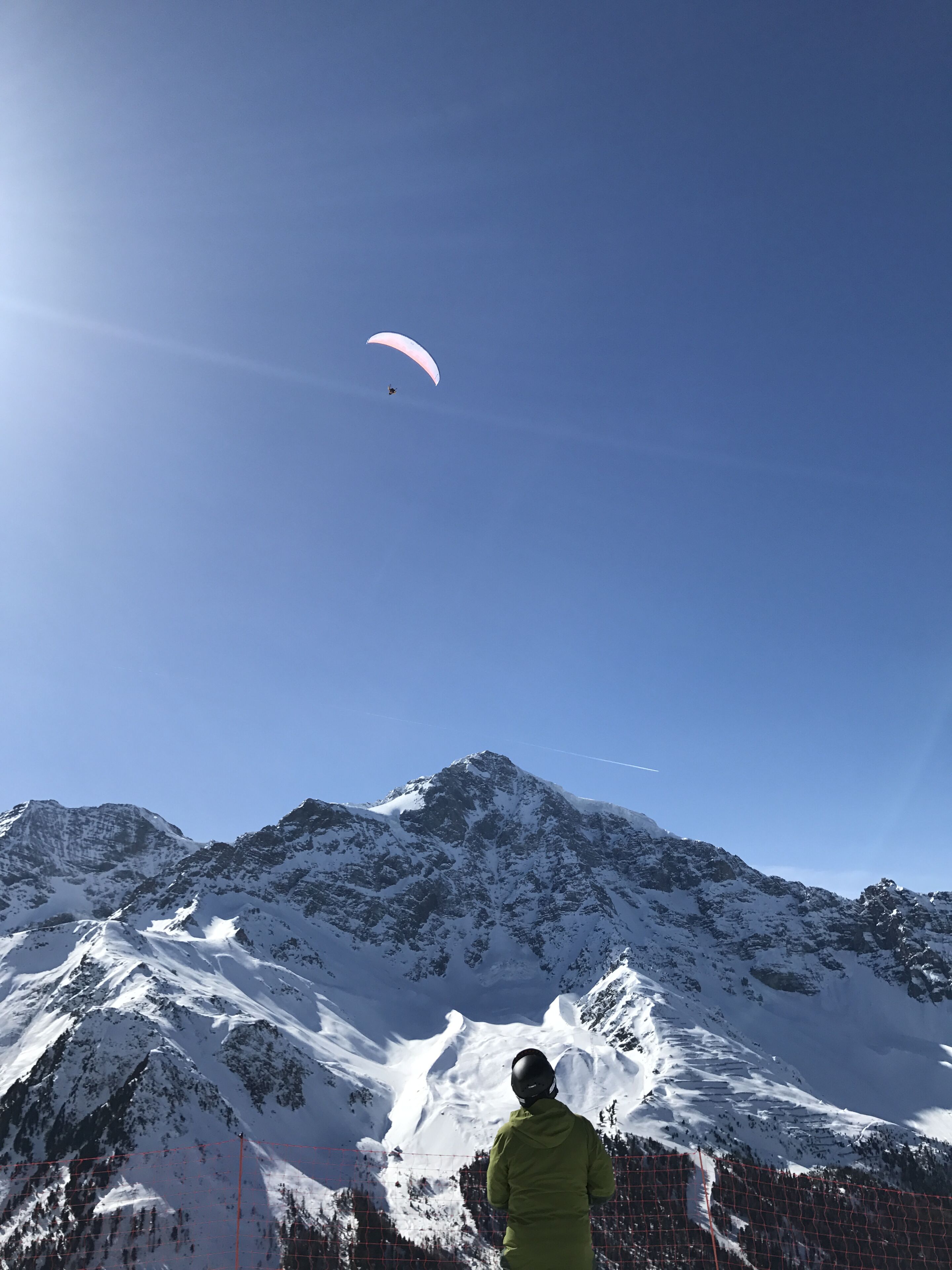 A person in a green jacket observing a paraglider in a mountainous area with snow-covered peaks.