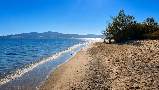 Una playa con agua cristalina, montañas lejanas y algunas personas sentadas en la arena.