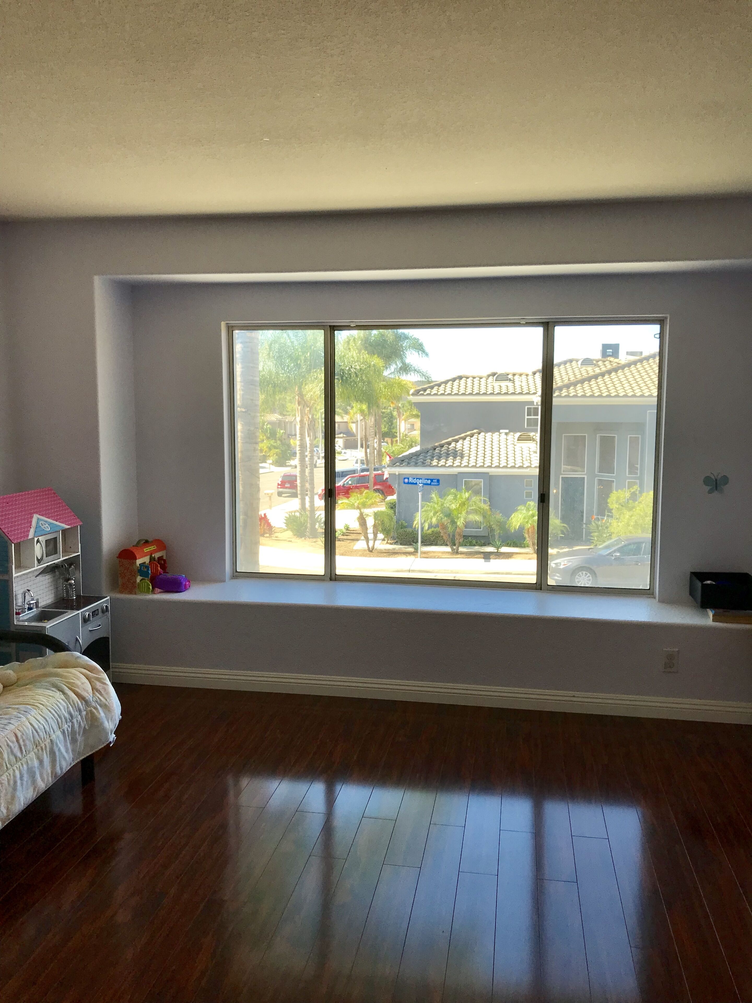 A room with a large window, wooden flooring, a bed, a small desk, and a toy house.