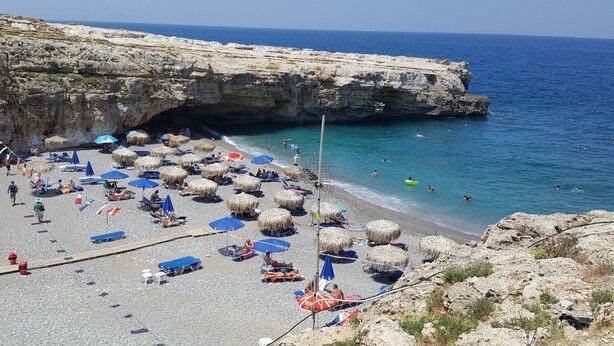 A beach with thatched umbrellas, sun loungers, and a rocky cliff.