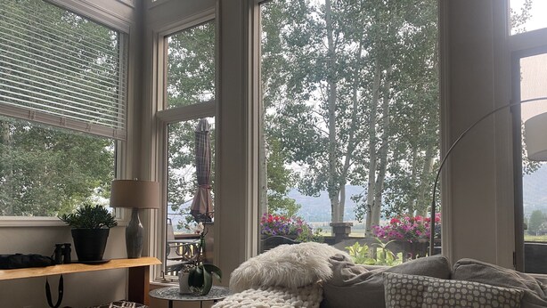 Un salon chaleureux avec une grande fenêtre, un canapé, une lampe et une vue sur les arbres et les fleurs à l’extérieur.