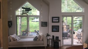 Un salon chaleureux avec une grande fenêtre, un canapé agrémenté de coussins à motifs, une petite table avec une plante, et un barbecue à l’extérieur.