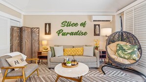 Een woonkamer met een bank, fauteuil en een ronde salontafel met een dienblad vol drankjes. Een decoratief bord aan de muur vermeldt “Slice of Paradise”.