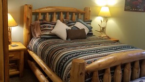 Een houten bed met een beddekleed met patroon en kussens, een houten hoofdbord, een nachtkastje met een lamp en een wandbord.