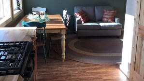 Une cuisine chaleureuse avec une table en bois, des chaises et une cuisinière à gaz. Un canapé gris et un tapis coloré se trouvent dans le salon.
