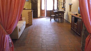 Un couloir carrelé mène à une salle à manger où se trouvent une table et des chaises. On y aperçoit également un meuble en bois et un téléviseur posé sur un meuble.