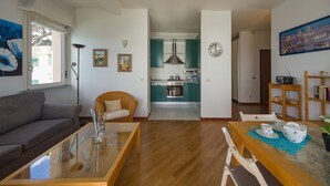 A modern living room with a sofa, coffee table, and a dining area with chairs and a table set for tea. A kitchen with teal cabinets and a stove is visible in the background. - Amazing seaview apartment Nervi - Genova (Genova)