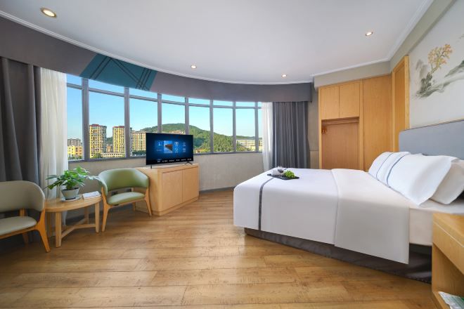 A hotel room with a bed, a desk, a TV, and a large window with a city view.