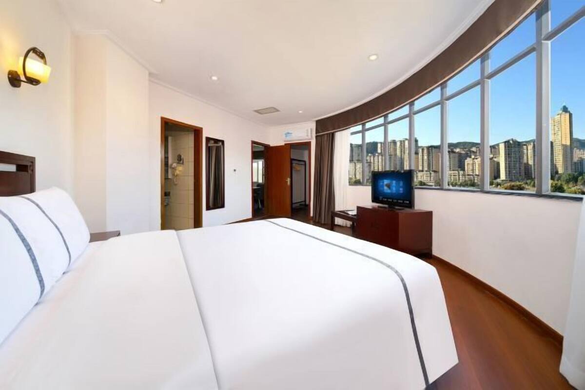 A hotel room with a large bed, a television, and a view of the cityscape through large windows.