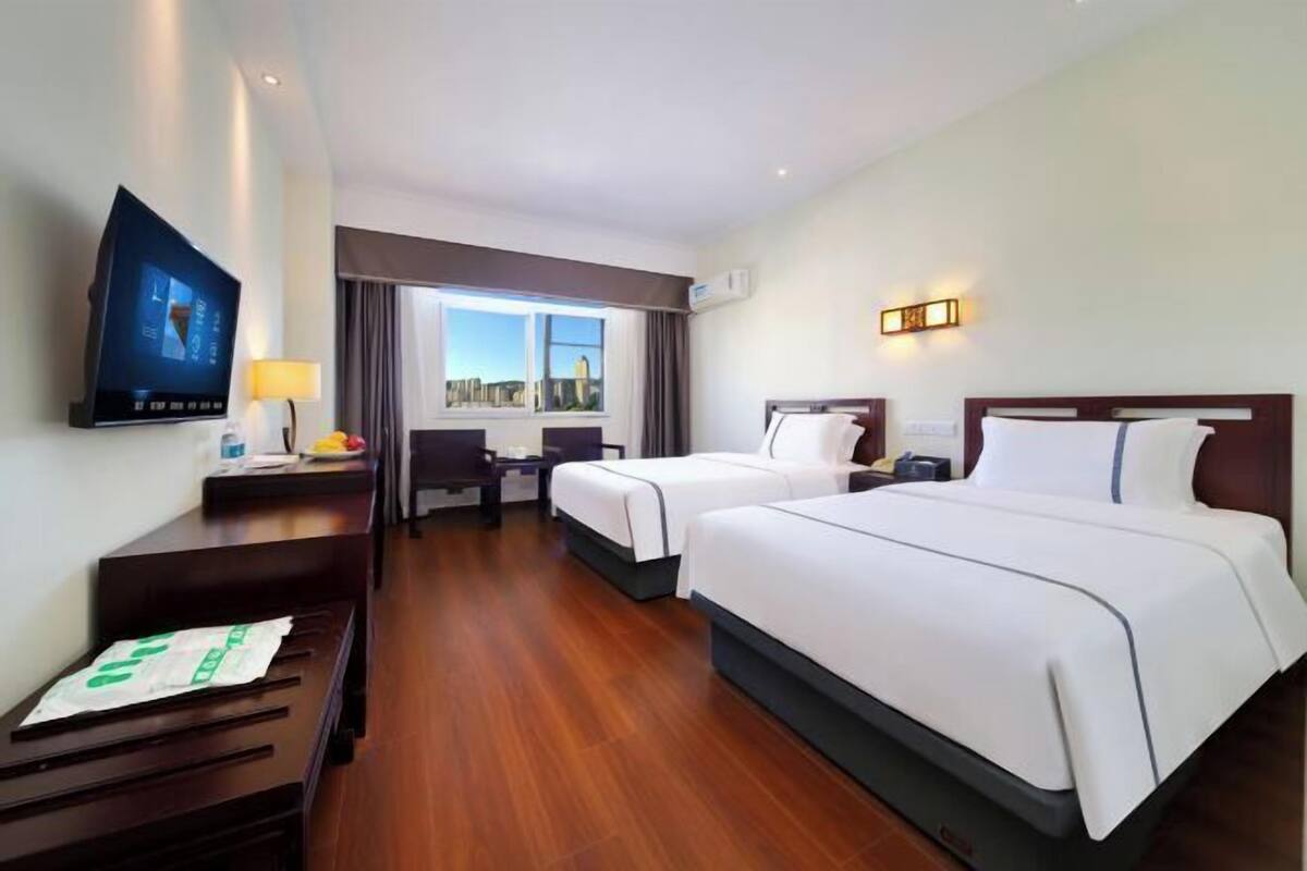 A hotel room with two beds, a TV, a desk, and a window with a city view.