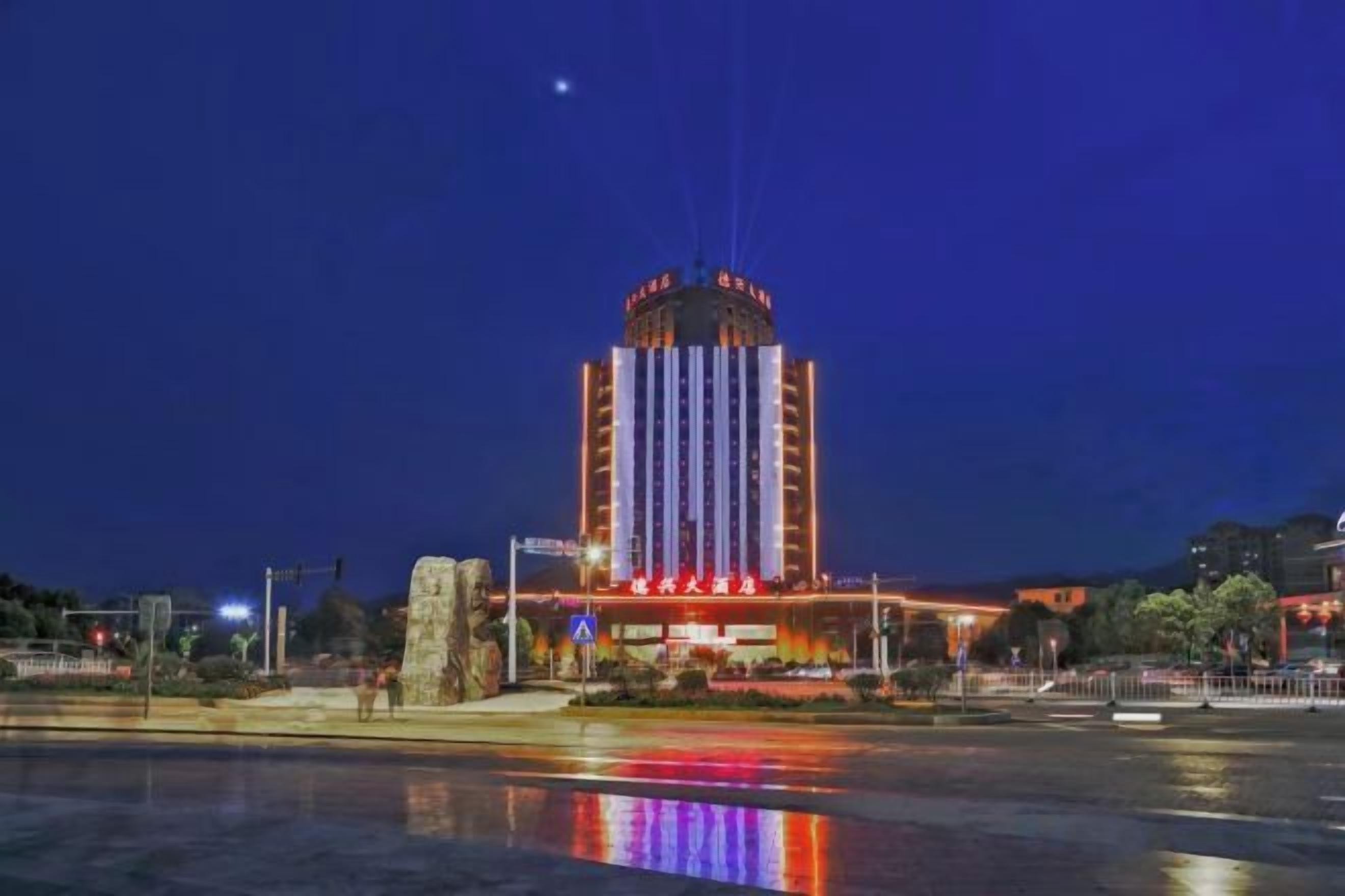 Un gran edificio con una torre central y varios niveles, ubicado junto a una masa de agua que refleja luces.