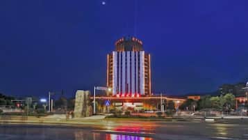 A large building with a central tower and multiple levels, situated next to a body of water reflecting lights.