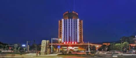 Un gran edificio con una torre central y varios niveles, ubicado junto a una masa de agua que refleja luces.