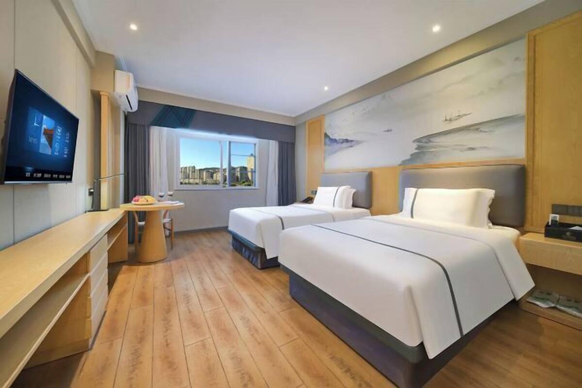 A hotel room with two beds, a desk, a TV, and a window with a city view.