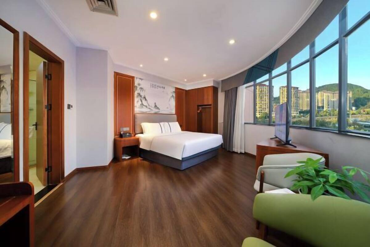 A hotel room with a bed, a desk, a chair, a large window with a view, and a wall with a painting.