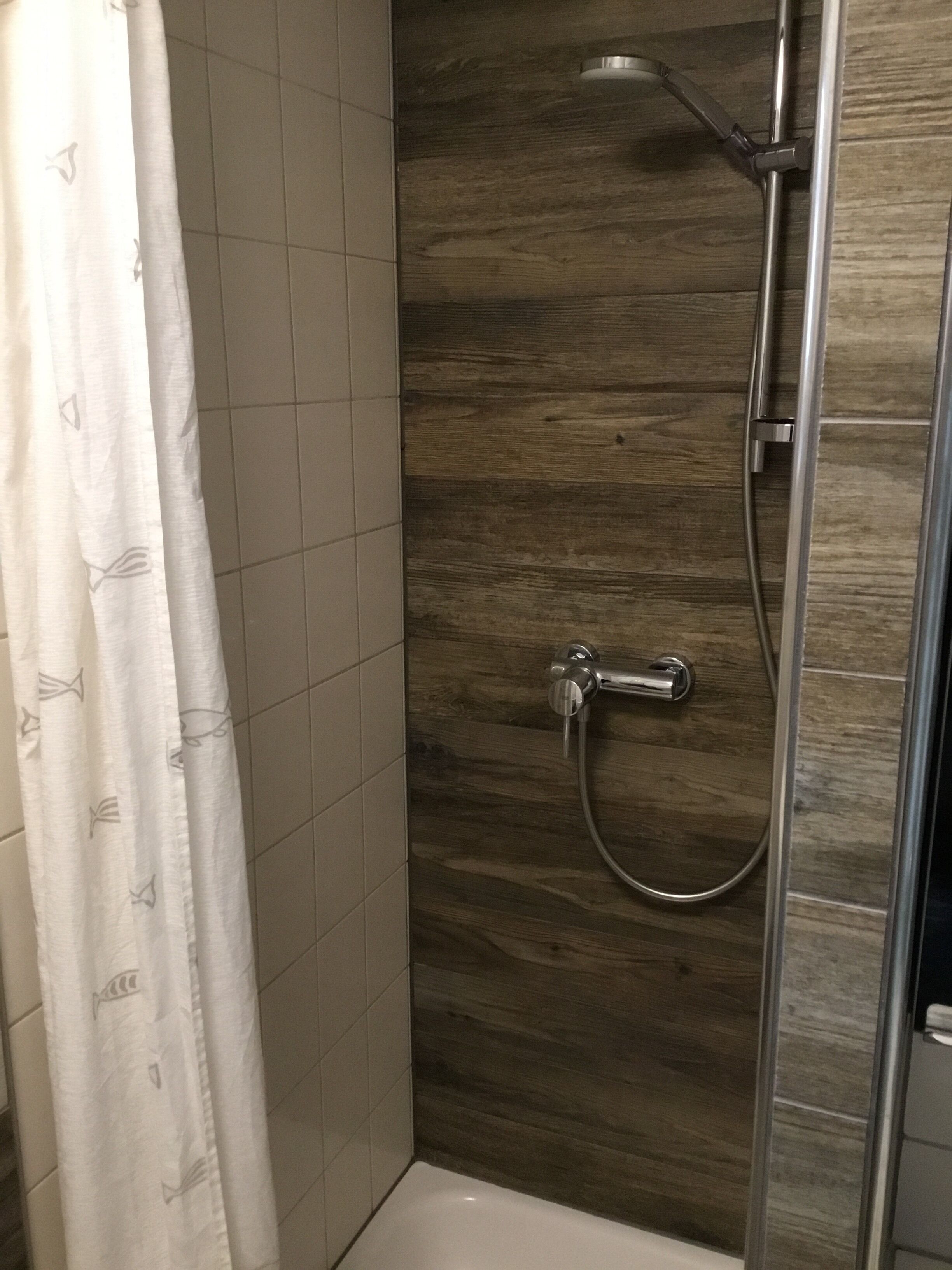 Eine Dusche mit Holzwand, Fliesenboden und weißer Duschvorhang mit Muster.
